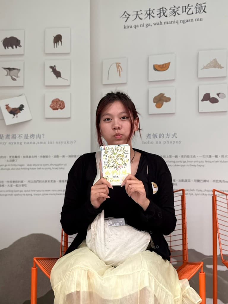 學生坐在「今天來我家吃飯」展牆前,拿著畫有小組人物的手寫卡片