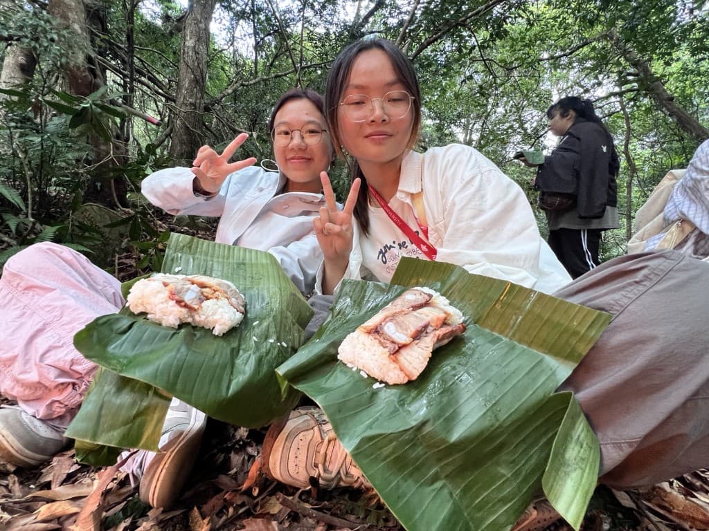 兩位學生在森林地面以香蕉葉盛裝餐食,向鏡頭比出和平手勢